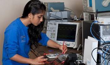 a person works on a circuit board with computers around her